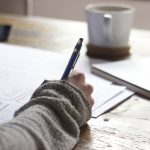 Picture of a woman writing on paper with a coffee cup in the background. Image used for blog about how to write a press release summary.