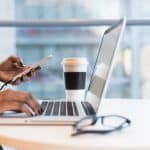 Picture of woman typing on computer, looking at phone, with coffee cup near by. Picture being used for blog, "Are Press Releases Driving Business Growth for CEOs?"