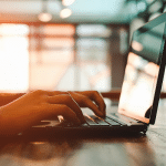 Picture of a person typing on a laptop. Computer is in the forefront, light in the background. Picture is being used for a blog post about how much does it cost to distribute a press release?