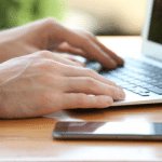 Image of a man's hands on the keyboard of a laptop. A smartphone is next to him. Image is being used for a Newswire blog title, Where do Press Releases Get Distributed?