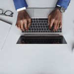 Flat lay image of an African American man in a blue suit typing on a Macbook. Image is being used for a blog about press release distribution outsourcing.