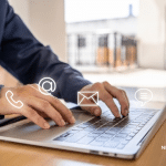 Picture of a man in a blue button down typing on a computer. There are four symbols near his hands, telephone symbol, ampersand, an envelope, and a speech bubble. Image is being used for a Newswire blog about a Media Database.