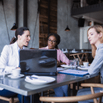 Picture of three business women in a coffee shop sitting in front of a computer and talking. Image is being use for a Newswire blog about Media Pitching improving the effectiveness of press release distribution.