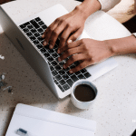 Picture of a woman's hands typing on a Macbook with a cup of coffee and notebook on the table. Image is being used for a Newswire blog post about the benefits of using a wire service.