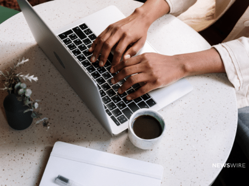 Picture of a woman's hands typing on a Macbook with a cup of coffee and notebook on the table. Image is being used for a Newswire blog post about the benefits of using a wire service.
