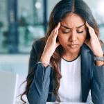 Picture of a business woman with dark complexion looking stressed and holding her hands at her temples. This image is being used for a blog about the use of media monitoring for crisis management.