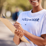 Picture of an African American woman holding a clipboard and smiling at a charity event. Her shirt says, "volunteer." This image is being used for a Newswire blog about How to Write a Nonprofit Press Release