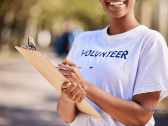 Picture of an African American woman holding a clipboard and smiling at a charity event. Her shirt says, "volunteer." This image is being used for a Newswire blog about How to Write a Nonprofit Press Release