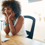 African American woman sitting in front of her computer with her head in her hands and a smile on her face.